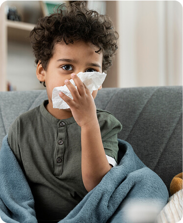 Child sitting on a sofa with a tissue, showing symptoms of cold and fever.